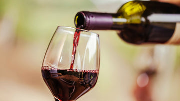 A person pouring red wine from a 750ml bottle into a stemless wine glass on a dinner table, with four other glasses already filled.